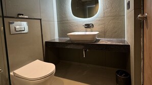 A modern bathroom with a toilet, a sink with a mirror above it, and a trash can. - Yuvraj Landmark (Mysore)