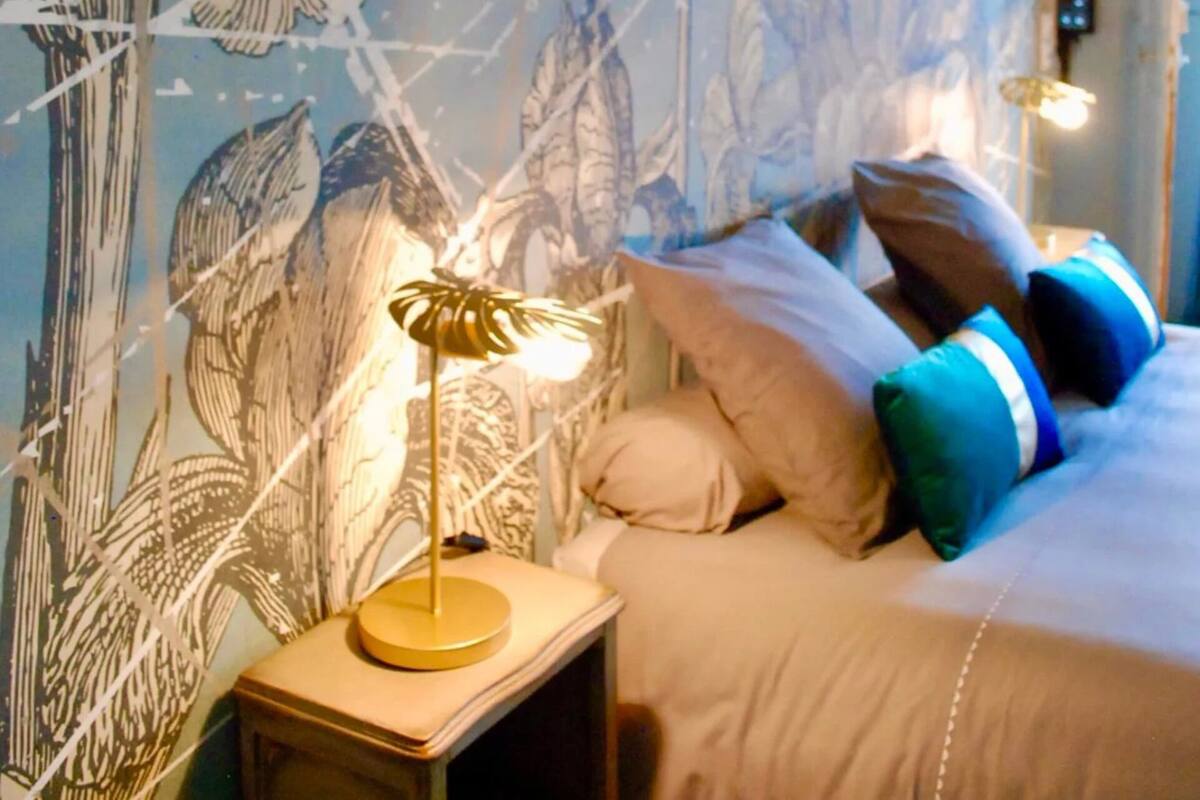 A bedroom with a bed, pillows, a bedside lamp, and a wall with a floral mural.