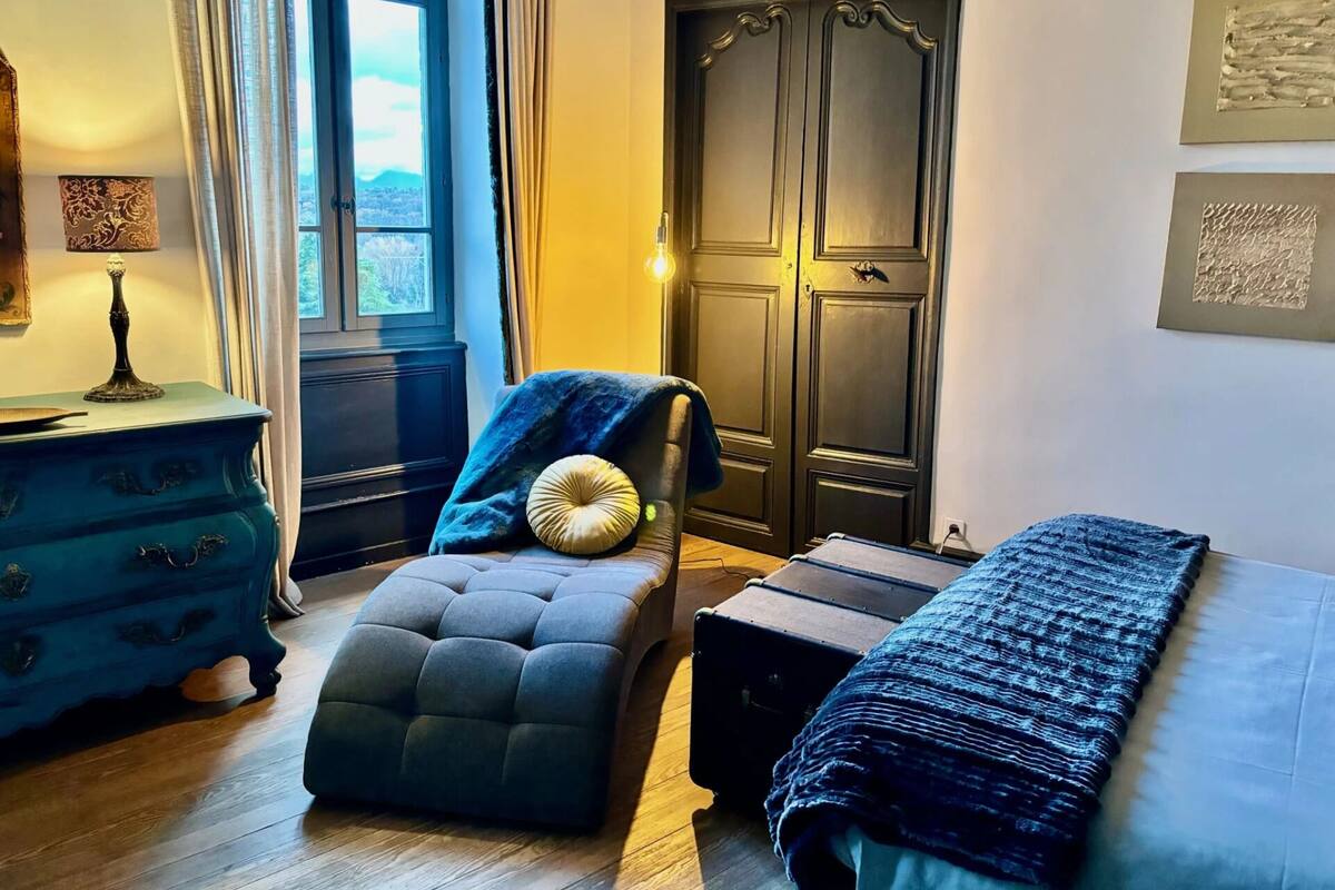 A bedroom with a large window, a dark wooden wardrobe, a blue upholstered chair, a bedside table, and a bed with a blue blanket.