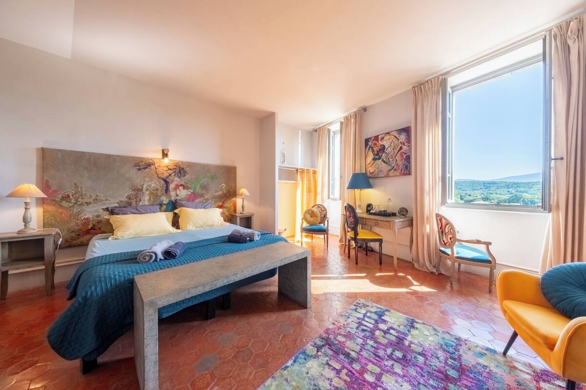 A hotel room with a bed, a desk, a chair, a large window, and artwork on the walls.