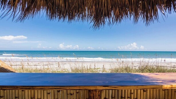 A beach view from a covered area with a bamboo fence and a blue table. - Welcome to “Salty Dog” - named after the old sea captain, this beautiful house directly on the Atlantic Ocean is decorated throughout with a nautical theme. With the breathtaking view of the ocean out your back door you will feel as though you are ac (Indialantic)