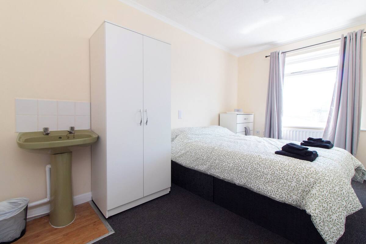 Een slaapkamer met een bed, een kledingkast, een wastafel en een raam met gordijnen.