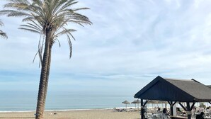 A beachside restaurant with outdoor seating, a palm tree, and the ocean in the background. - Golden Sunrise – Santa Clara – direct access to the beach (Torremolinos)