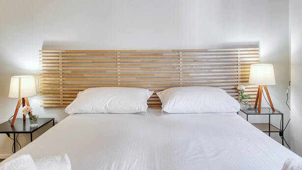 A neatly made bed with white linens, a wooden headboard, two bedside lamps, and a small vase with flowers on each side.