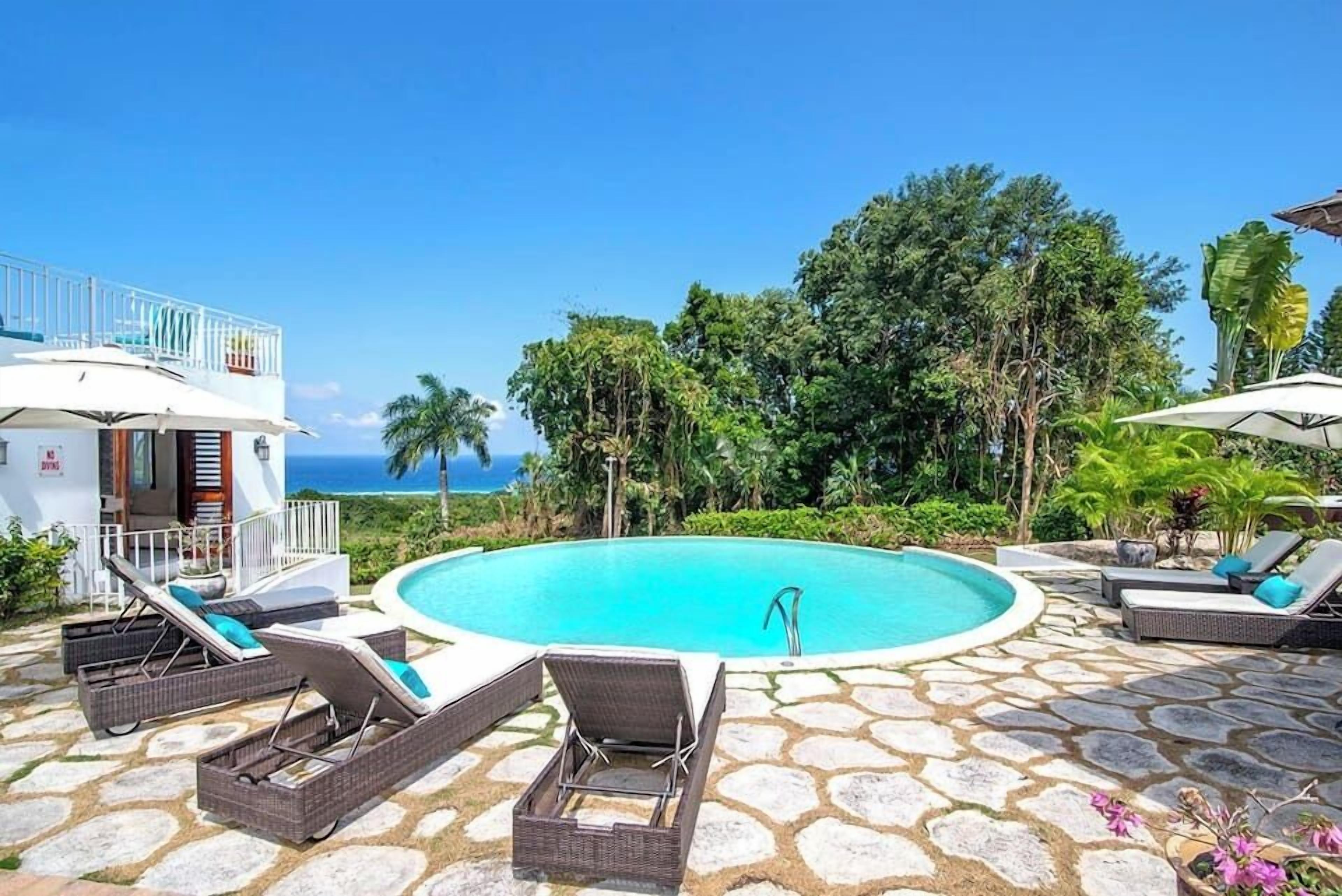 A pool area with lounge chairs, a stone patio, and a view of the ocean.