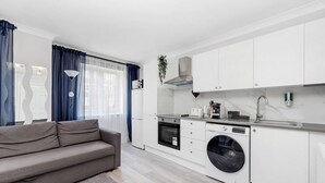 Una cocina compacta con horno integrado, microondas y lavadora, un sofá gris y un ventanal con cortinas.