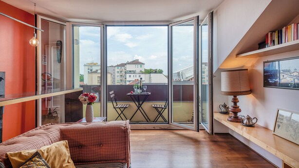 Een moderne woonkamer met een grote glazen deur naar een balkon, een bank, een klein tafeltje met een vaas, en een boekenkast met boeken.