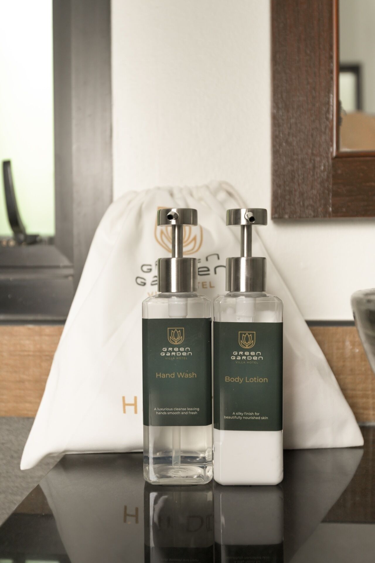 Two bottles of Green Garden Villa Hotel hand wash and body lotion on a reflective surface with a white bag in the background.