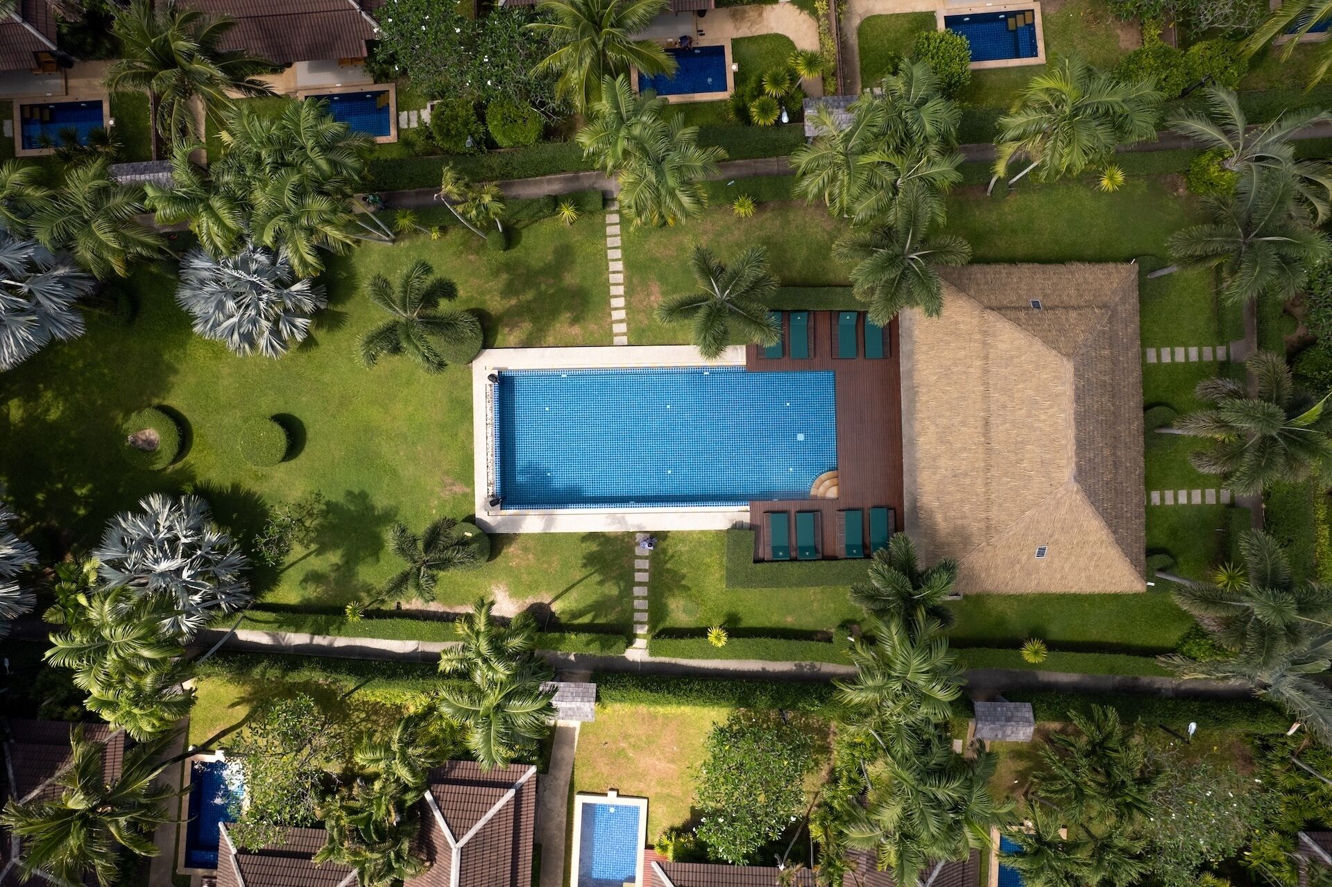 An aerial view of a resort with a central swimming pool, surrounded by palm trees and a well-maintained lawn.