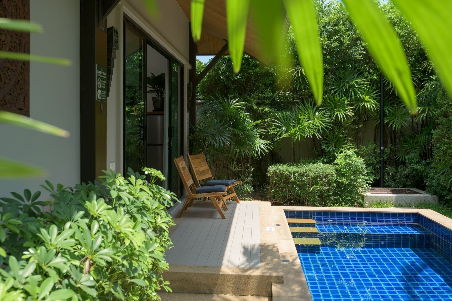 A pool area with a wooden deck, a lounge chair, and lush greenery.