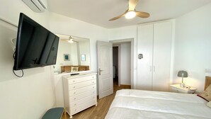 Quarto com cama, penteadeira, TV fixada na parede, ventilador de teto e mesa de cabeceira com abajur.