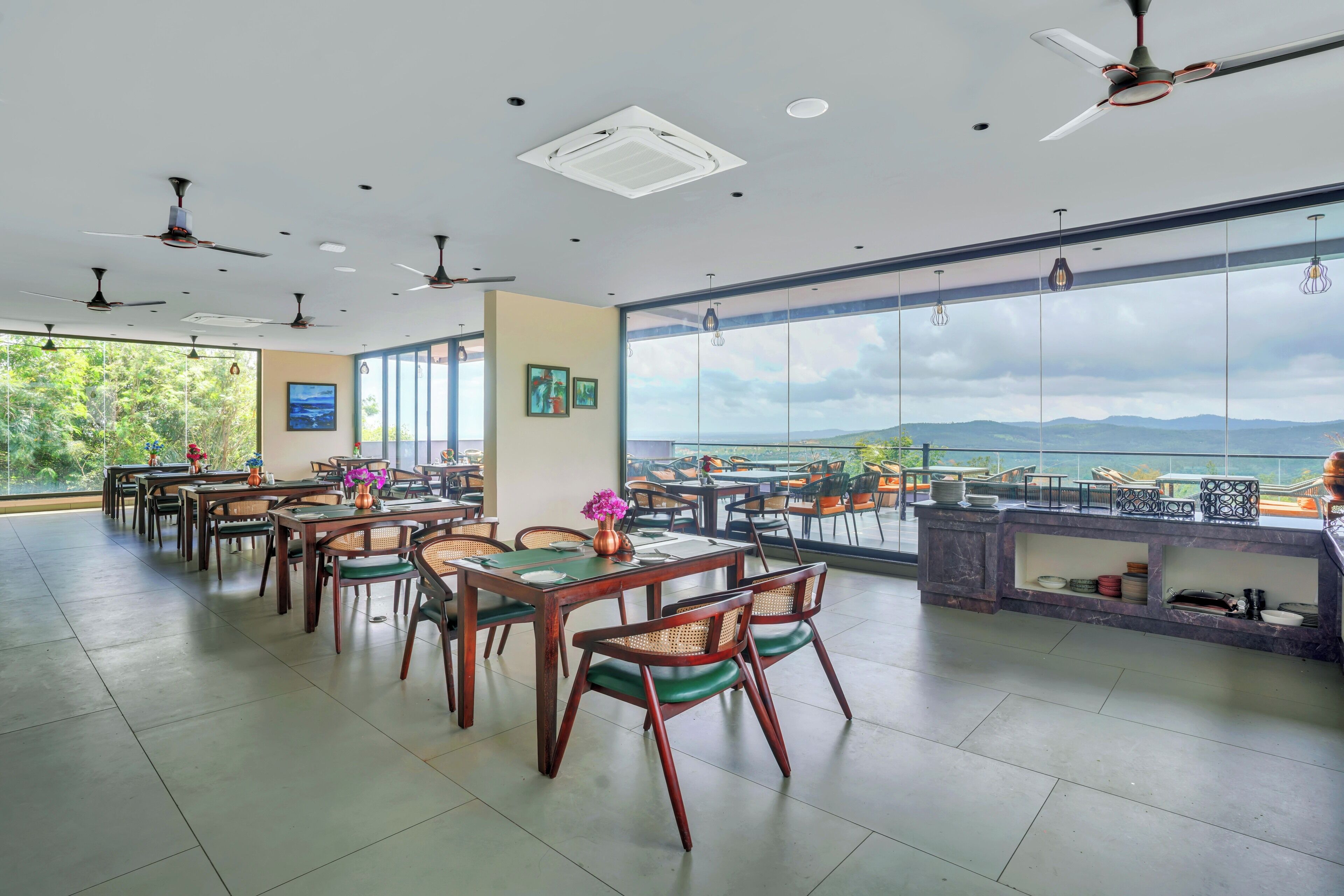 A spacious dining area with multiple tables and chairs, ceiling fans, and large windows offering a view of the outdoors.