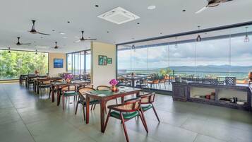 A spacious dining area with multiple tables and chairs, ceiling fans, and large windows offering a view of the outdoors.