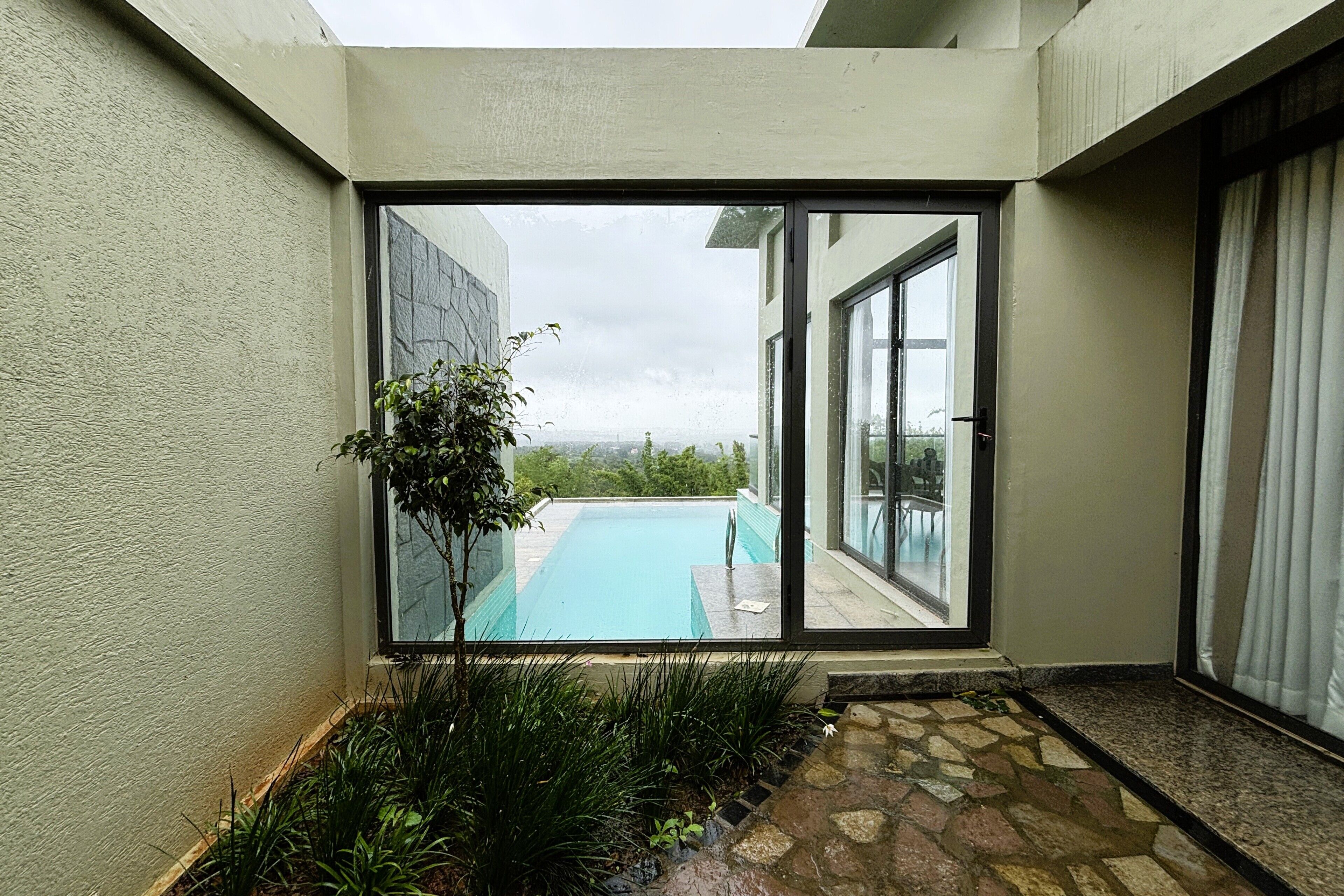 A view from a room with a glass door leading to a swimming pool, a stone pathway, and a small garden area.