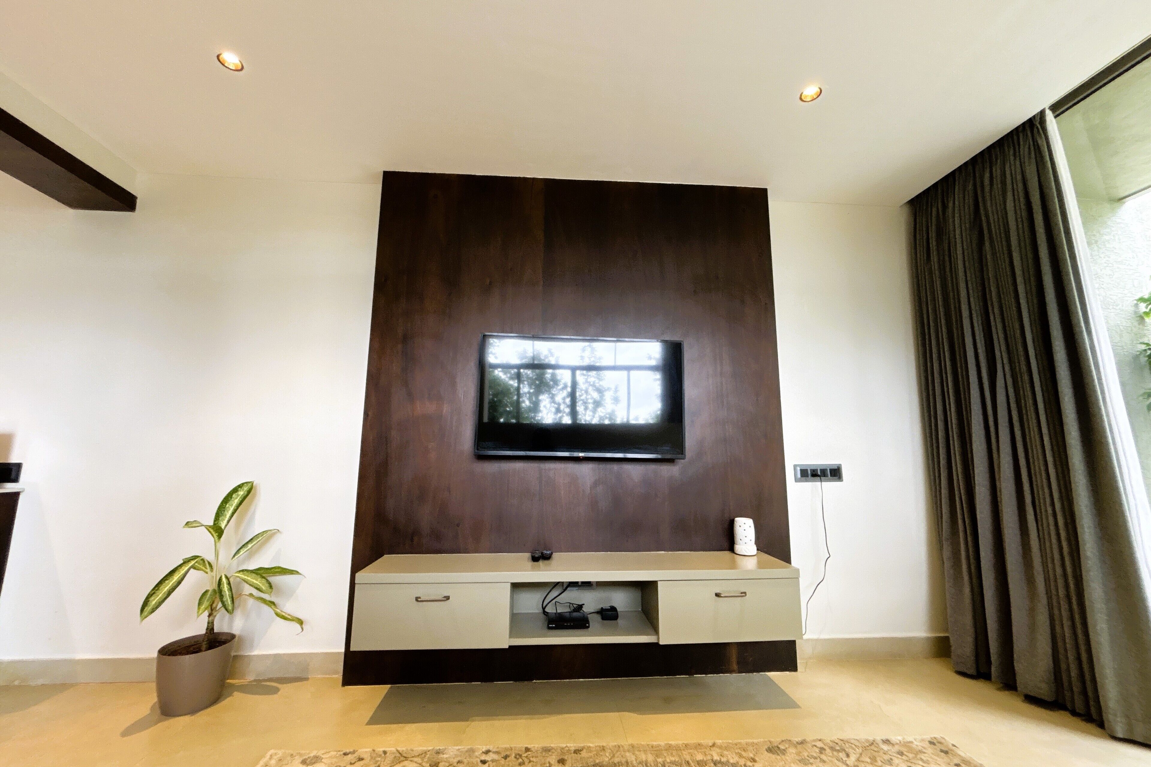 A modern living room with a flat-screen TV mounted on a dark wooden wall, a built-in entertainment unit below, a potted plant, and a large window with curtains.