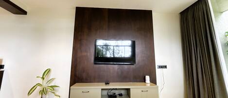 A modern living room with a flat-screen TV mounted on a dark wooden wall, a built-in entertainment unit below, a potted plant, and a large window with curtains.