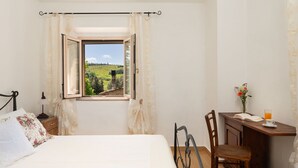 A bedroom with a bed, a desk, a chair, a window with a view of a vineyard, and a vase with flowers on the desk.
