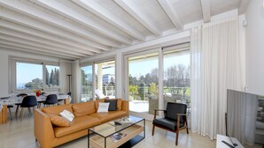 Una sala moderna con sofá, mesa de centro y sillas. Hay una zona de comedor con mesa y sillas. Amplias ventanas ofrecen vistas al exterior.