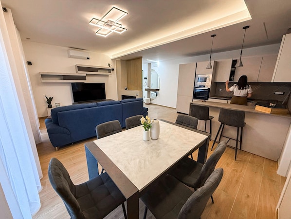 A modern living room with a blue sofa, a dining table with chairs, and a kitchen area with a person working at the counter.