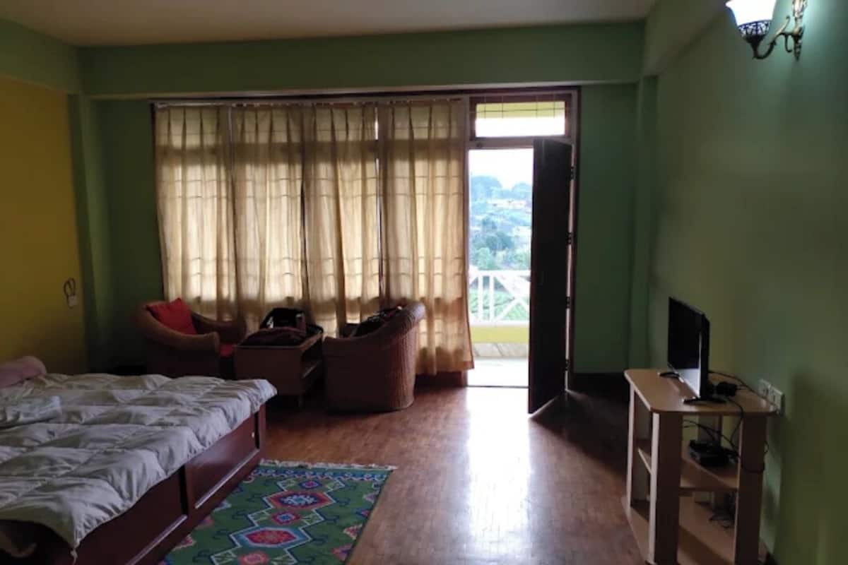 Ein Schlafzimmer mit Bett, Schreibtisch mit Computer, Stuhl, Fenster mit Vorhängen und Blick ins Freie.