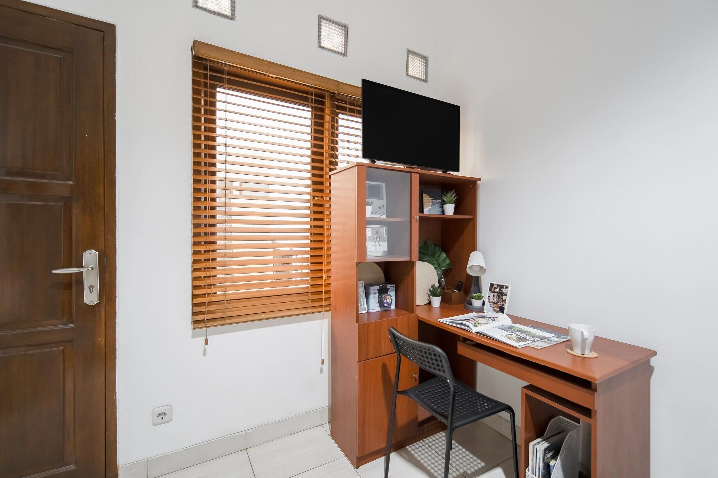 Una habitación con un escritorio de madera, una silla, un televisor montado en la pared y una ventana con persianas.