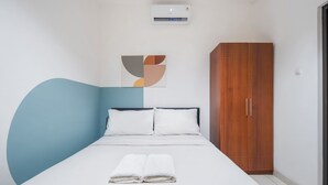 A bedroom with a bed, white pillows, a blue accent wall, a wooden wardrobe, and a wall-mounted air conditioner.