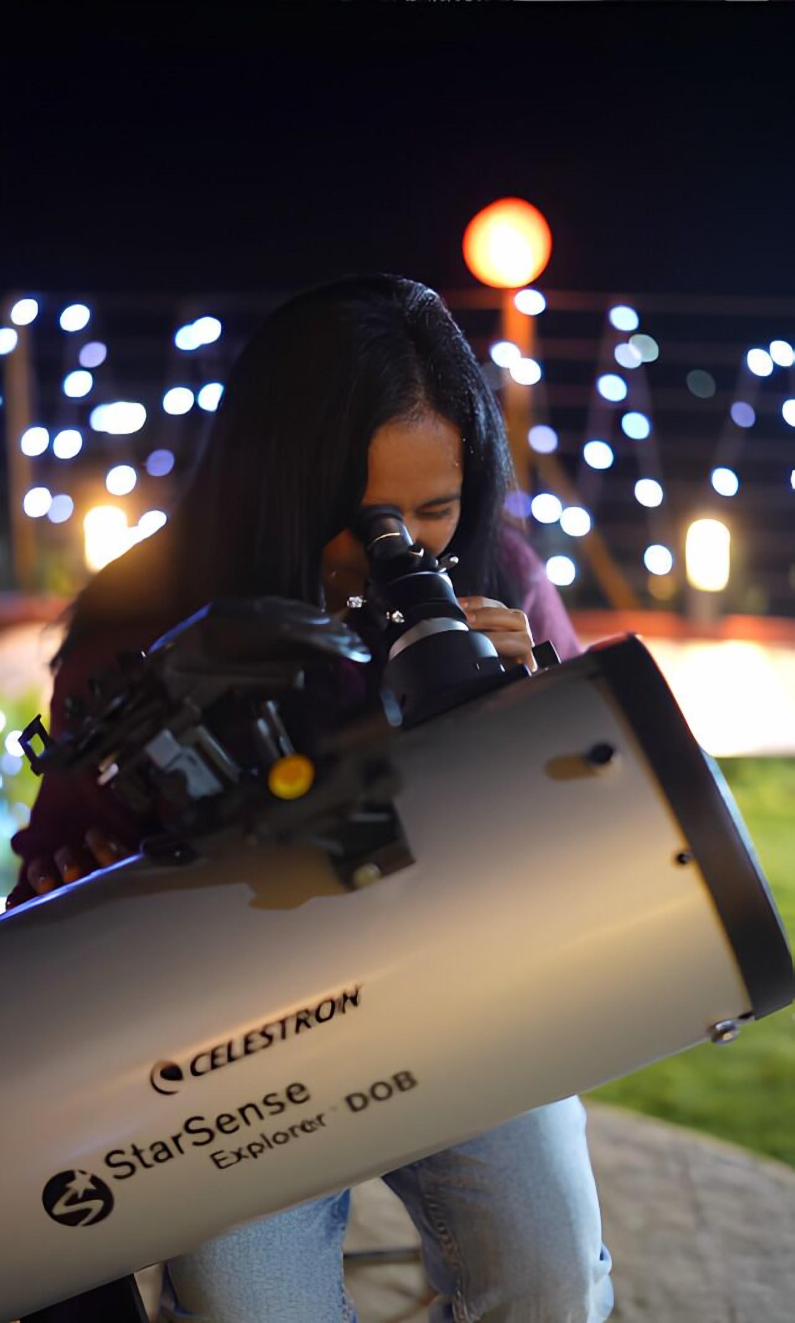 A person using a telescope labeled "StarSense Explorer - DOB" outdoors at night.