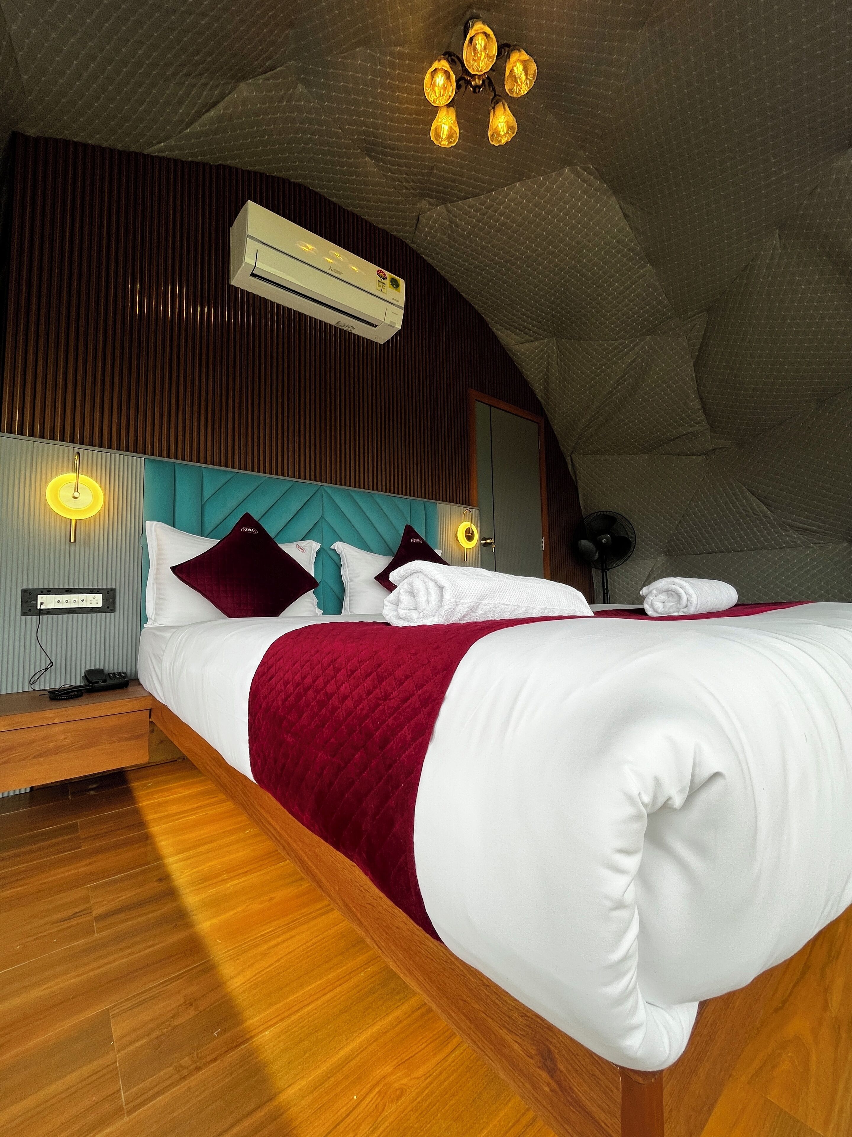 Un dormitorio moderno con suelo de madera, una cama grande con ropa de cama blanca y burdeos, un cabecero verde azulado y un diseño de techo único.