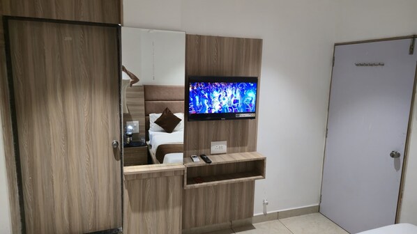 Una habitación de hotel con un armario de madera, una cama con ropa de cama blanca, un televisor montado y una mesita de noche con un control remoto y un libro.