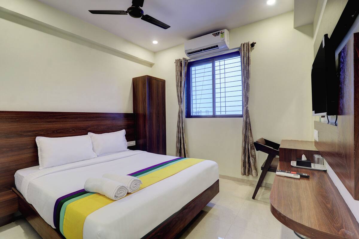 A hotel room with a bed, a desk, a TV, and an air conditioning unit.