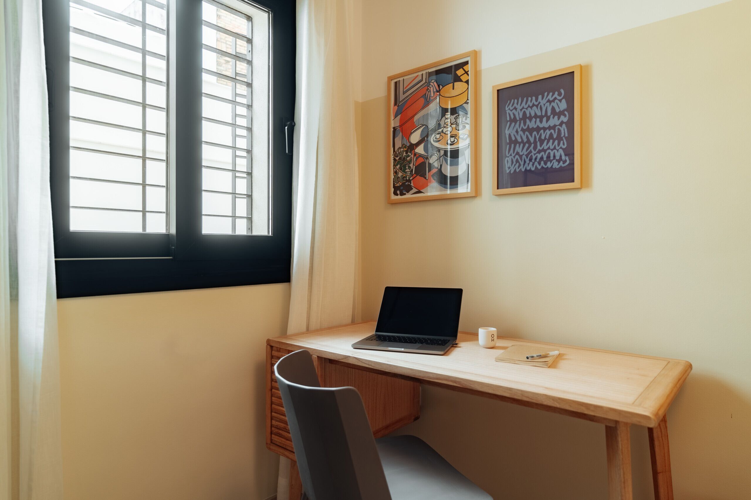 A room with a wooden desk, a laptop, a chair, a window with blinds, and two framed artworks on the wall.