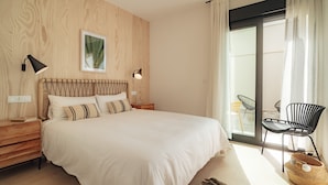 Un dormitorio con una cama grande, cabecera de madera, dos mesitas de noche, una lámpara de pared, un cuadro enmarcado y vistas al balcón exterior.