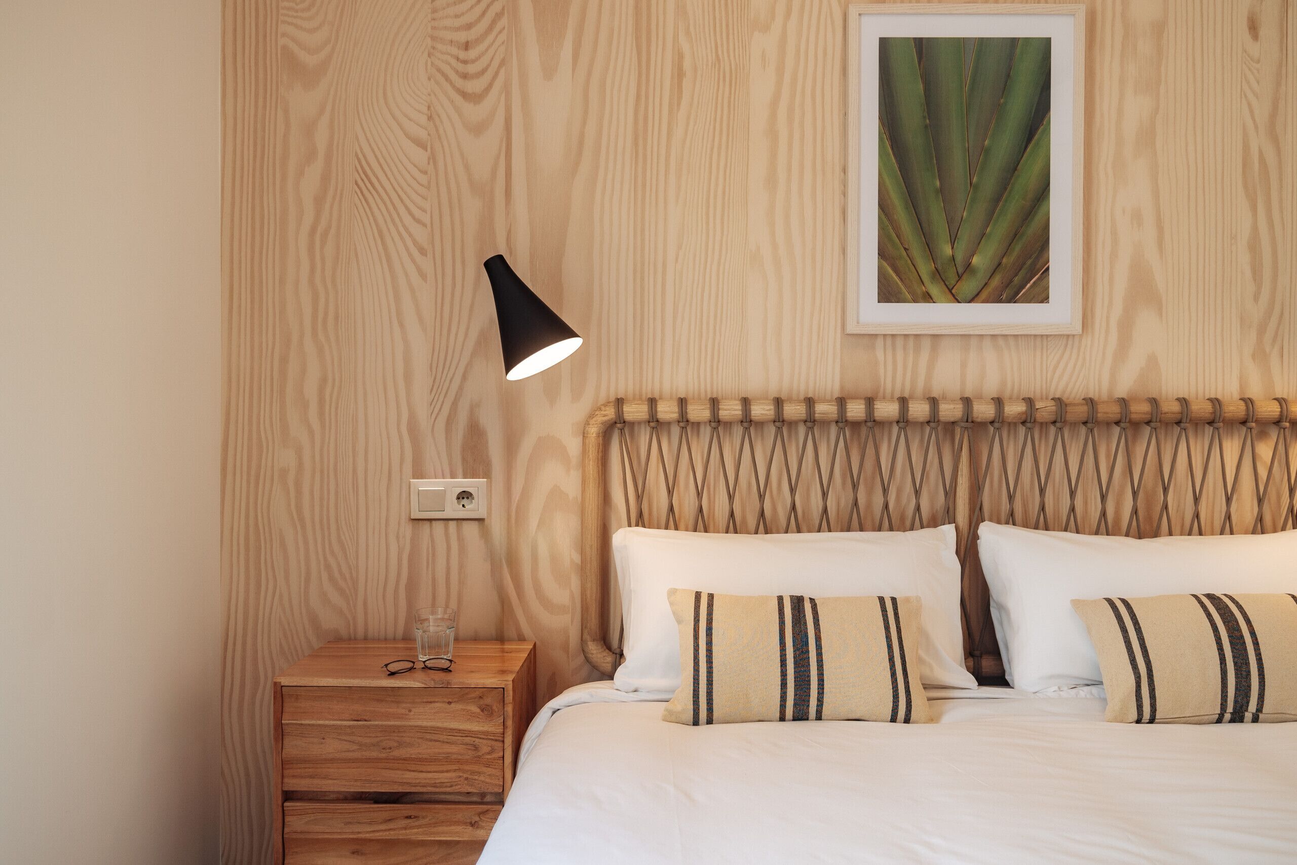A neatly arranged bedroom with a wooden headboard, a bedside table with a lamp, and a framed plant on the wall.