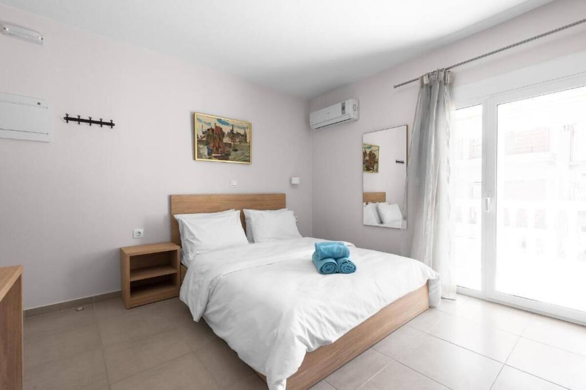 Una habitación de hotel moderna con una cama grande, una mesita de noche de madera, un estante pequeño y un ventanal corredero al exterior.