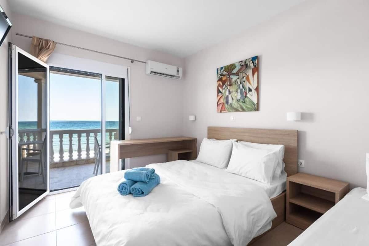 Habitación de hotel con cama, mesita de noche, un cuadro y puerta corredera a un balcón con vistas al mar.