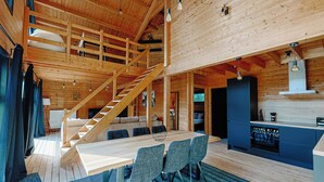 A spacious wooden cabin with a large dining table, multiple chairs, a kitchen area, and a staircase leading to an upper level. - Chalet "Les Alpages (Durbuy)