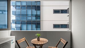 Ein Balkon mit rundem Holztisch und zwei Holzstühlen, einer Topfpflanze und Blick auf ein modernes Gebäude mit großen Fenstern.