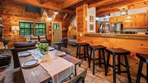 A cozy log cabin interior with a dining area, bar stools, a kitchen with wooden cabinets, and a staircase leading to another level. - Dog OK | Hot Tub + Game Rm | Dollywood (Pigeon Forge)
