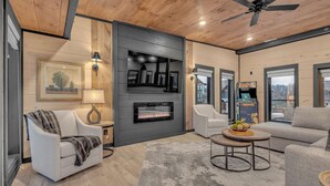 A modern living room with a fireplace, a ceiling fan, and a large window. - 10BR Luxury | 2 Cabins | Rooftop Lounges | Indoor Pools | Hot Tubs | Walk to Water Park | Sleeps 44 (Sevierville)
