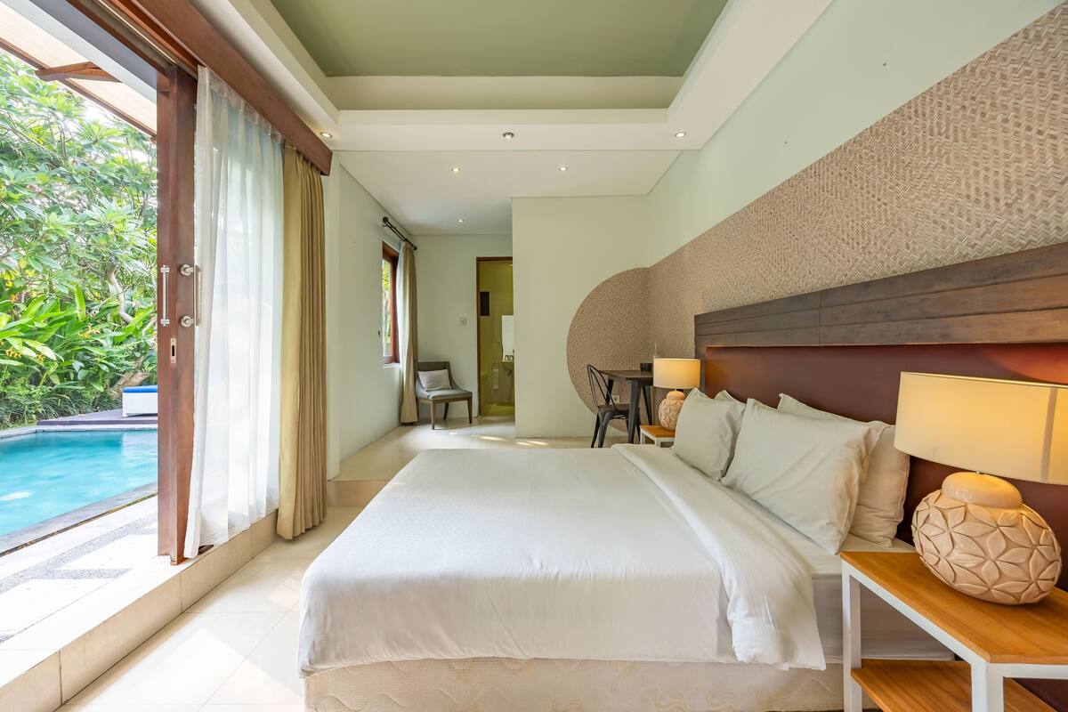 A spacious bedroom with a large bed, a wooden nightstand, and a view of a swimming pool.