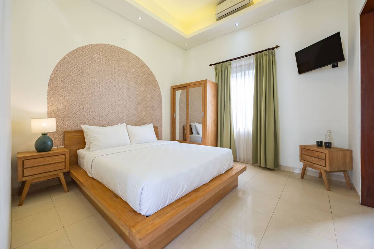 A modern hotel room with a large bed, wooden nightstands, a mirror, a television, and a small table with a bottle.