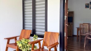 Une pièce avec un plancher en bois, deux chaises en bois, une petite table et une plante en pot. Il y a une porte avec une pancarte indiquant « KARLAD A1 ».