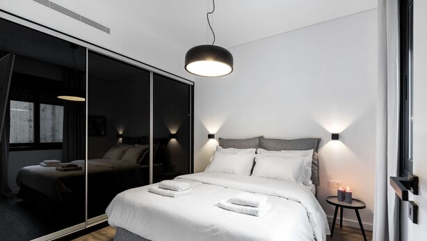 Een moderne slaapkamer met een grote spiegel, een bed met witte lakens, een ronde hanglamp en een klein nachtkastje met kaarsen.