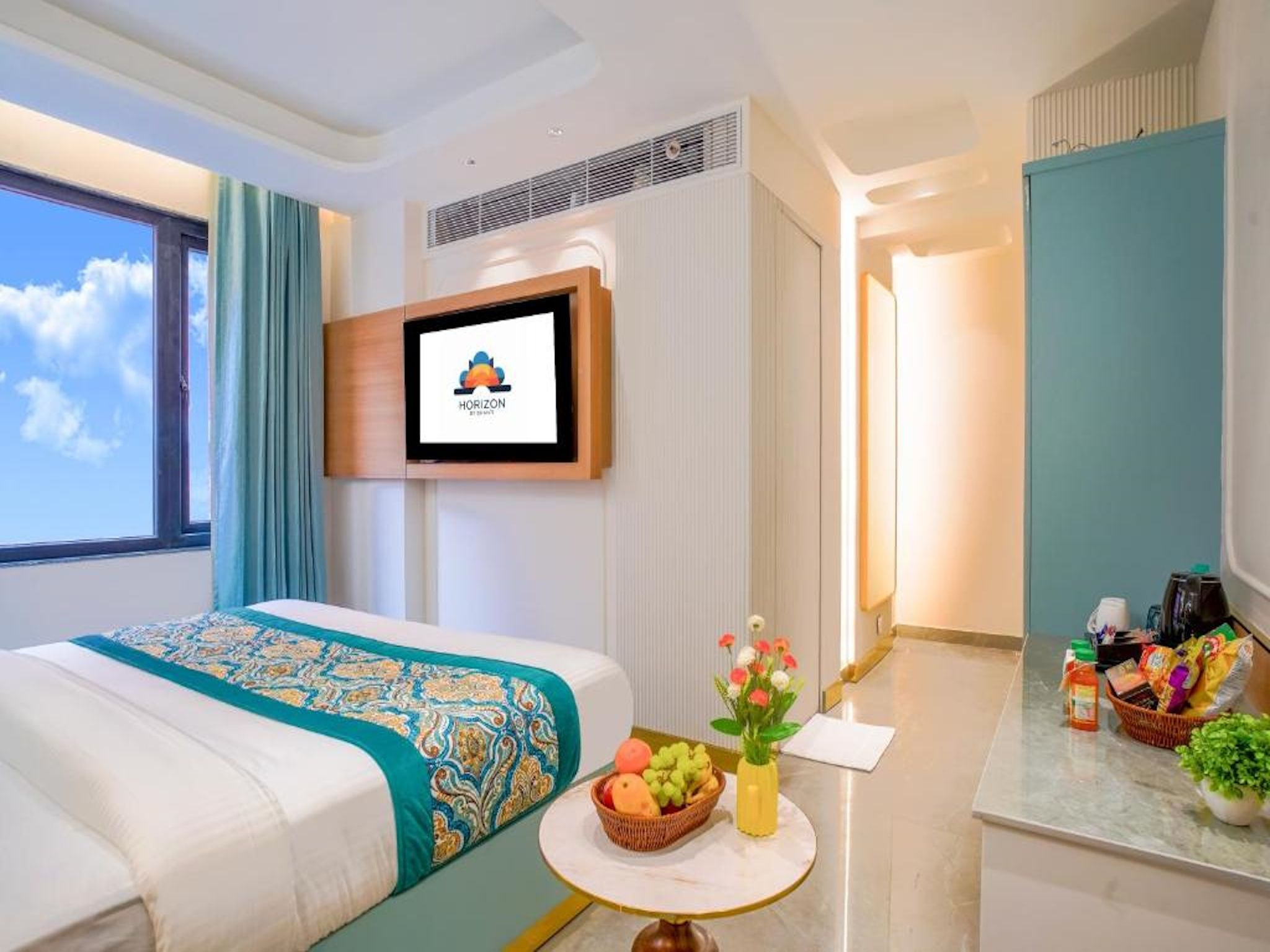 A hotel room with a bed, a television mounted on the wall, a small table with a fruit bowl, and a bathroom with a mirror and toiletries.