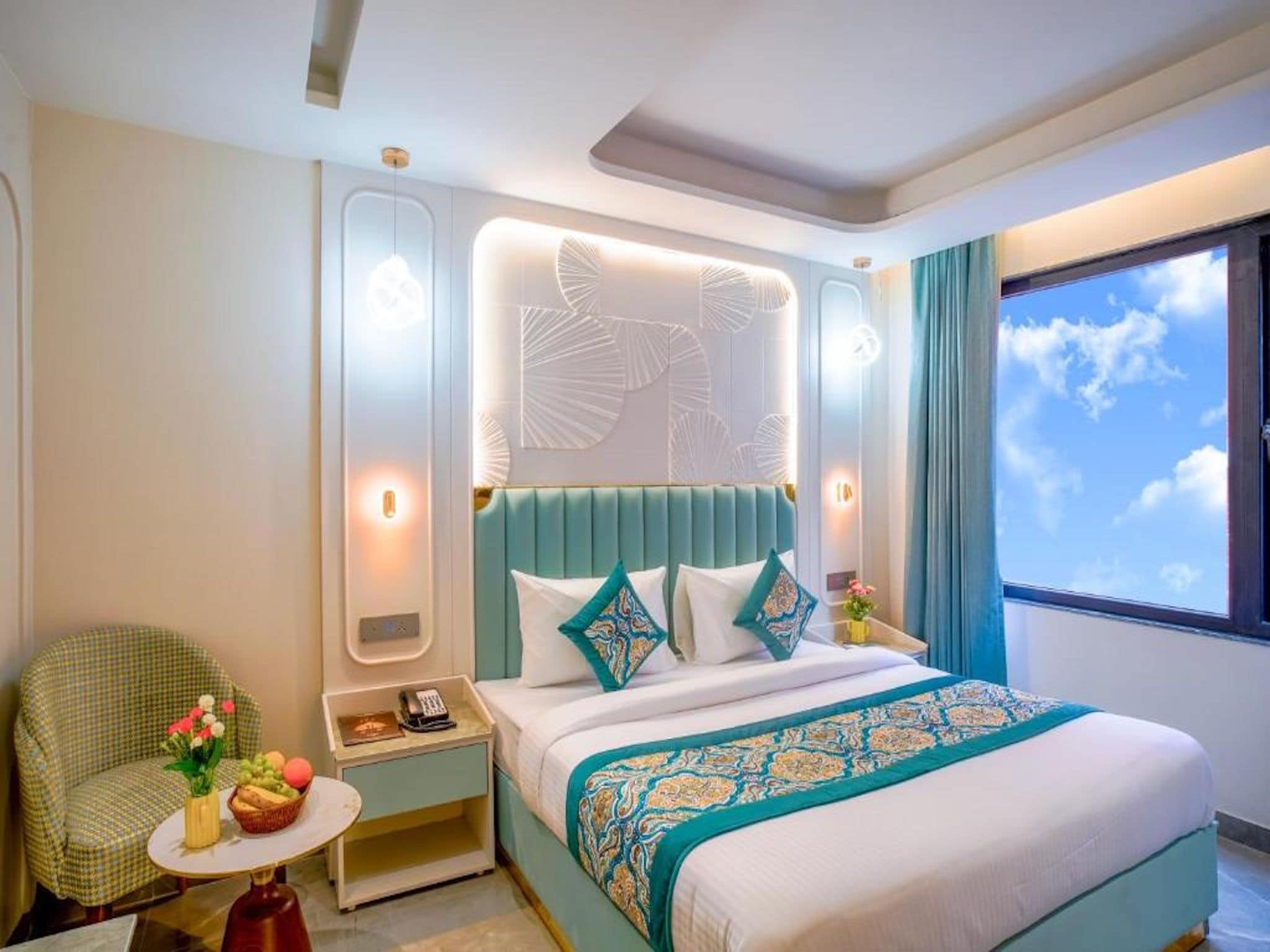 A hotel room with a bed, a nightstand, a chair, a small table with fruit, and a window with a view of the sky.