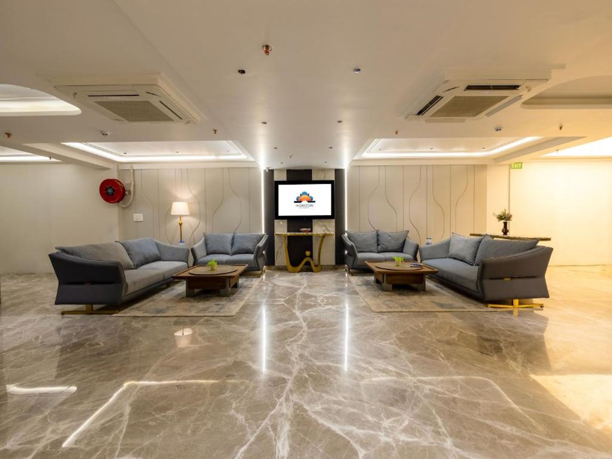 A spacious room with two sofas facing a television screen, a red fire alarm, and a marble floor.
