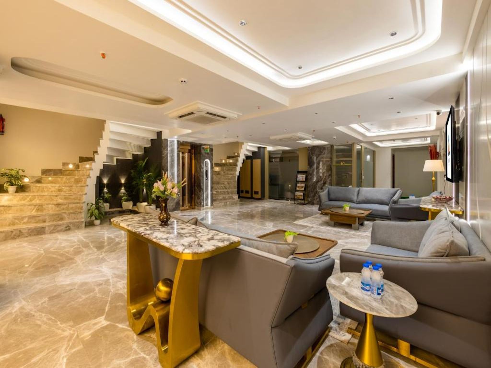 A spacious lobby with a marble floor, a central seating area with sofas and a coffee table, and a staircase with a modern design.