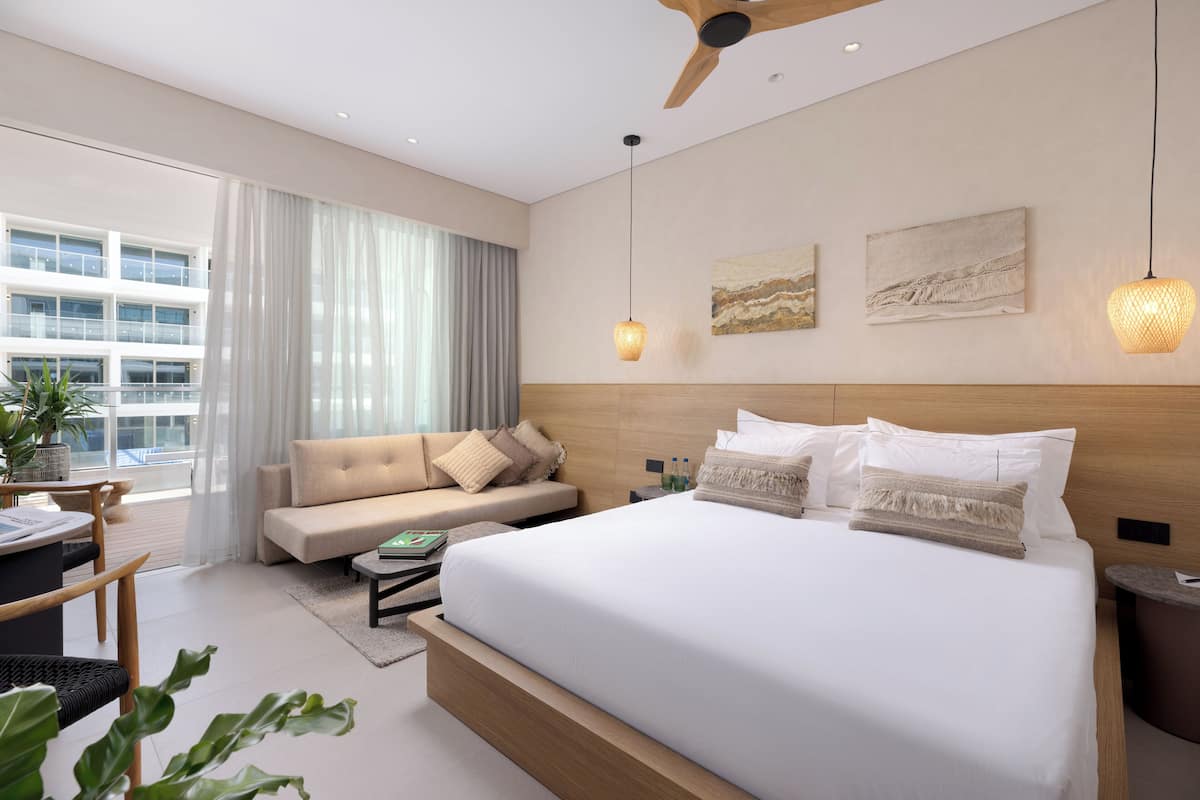 Una habitación de hotel moderna con una cama grande, un sofá, un comedor y vistas a un edificio a través de ventanas amplias.