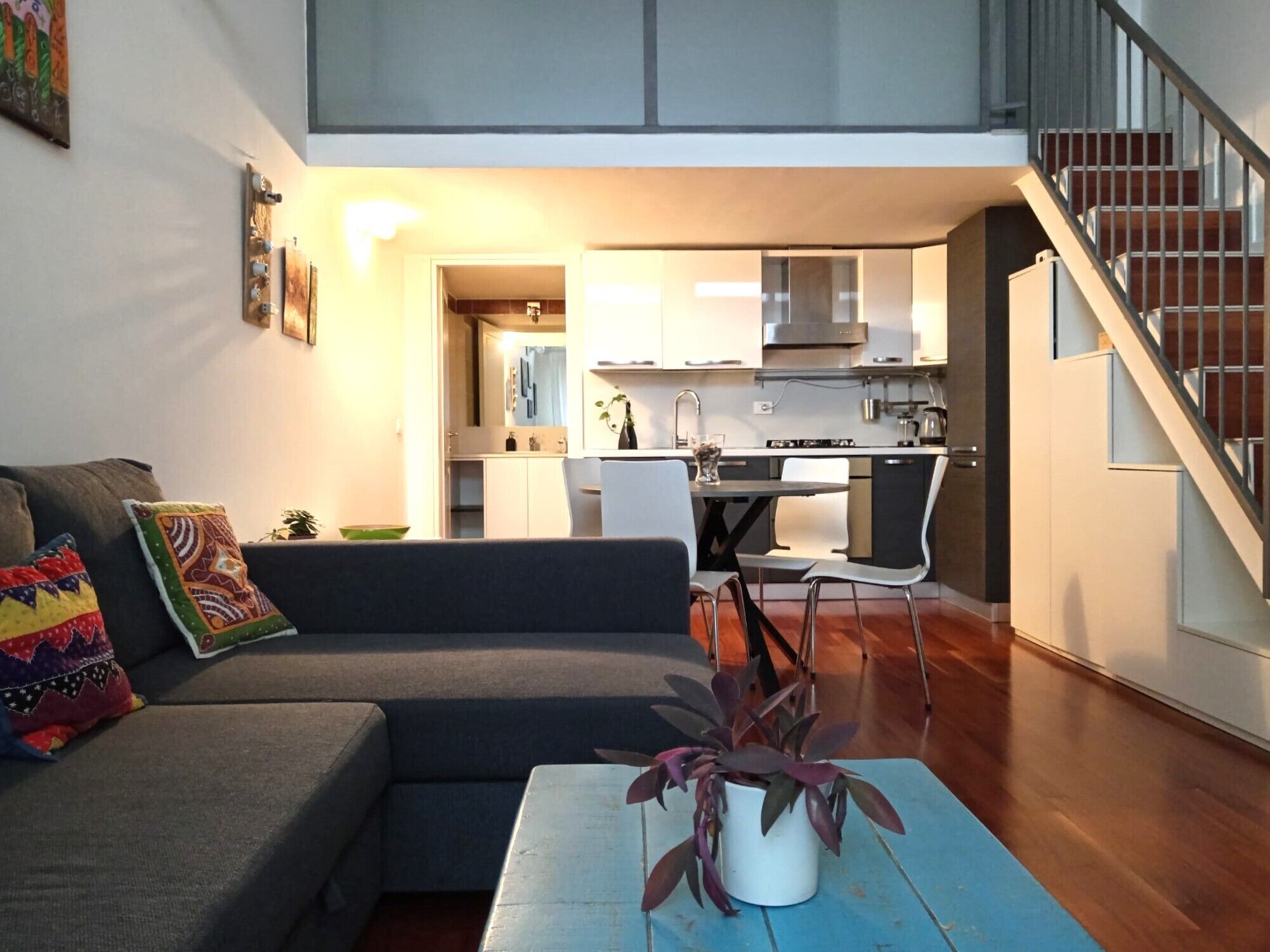 A modern living room with a grey sofa, colorful cushions, a blue coffee table with a potted plant, and a staircase leading to a kitchen area.