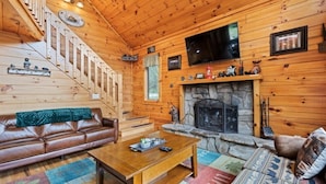 Ein Holzblockhaus-Interieur mit Kamin, einer Couch, einem Couchtisch, einem Fernseher, der über dem Kamin angebracht ist, und einer Treppe, die zum Obergeschoss führt.